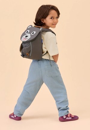 Child with brown hair wearing light pants and purple shoes, looking back while walking, carrying a gray backpack with a bear face.