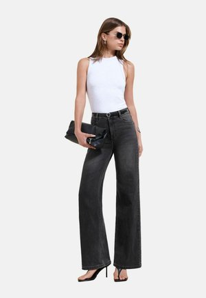 Kvinde iført en hvid ærmeløs top, sorte vide jeans, sorte hæle, solbriller og med en sort clutch står vendt mod venstre.