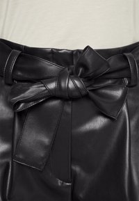 Zbliżenie na czarne skórzane spodnie z wysokim stanem i wiązanym paskiem w kokardę na tle jasnego topu.