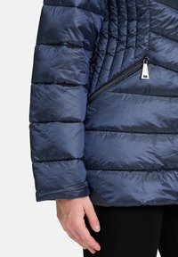Navy blauwe pufferjack met een gequiltte structuur, horizontale secties en een zijzak met een zilveren ritshendel.