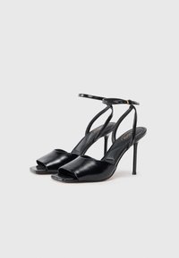 Sandalias de tacón alto de piel negra con punta abierta, correa delgada en el tobillo y un acabado suave. Presentan un tacón de aguja y un diseño minimalista.