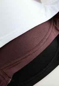 Trois t-shirts en coton pliés empilés, en blanc, mauve foncé et noir, montrant les ourlets cousus et la texture du tissu.