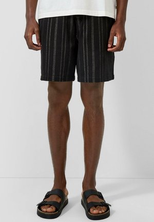 Mann trägt schwarze gestreifte Shorts, weißes Hemd und schwarze Sandalen mit Schnalle, steht vor einem einfarbigen hellgrauen Hintergrund.