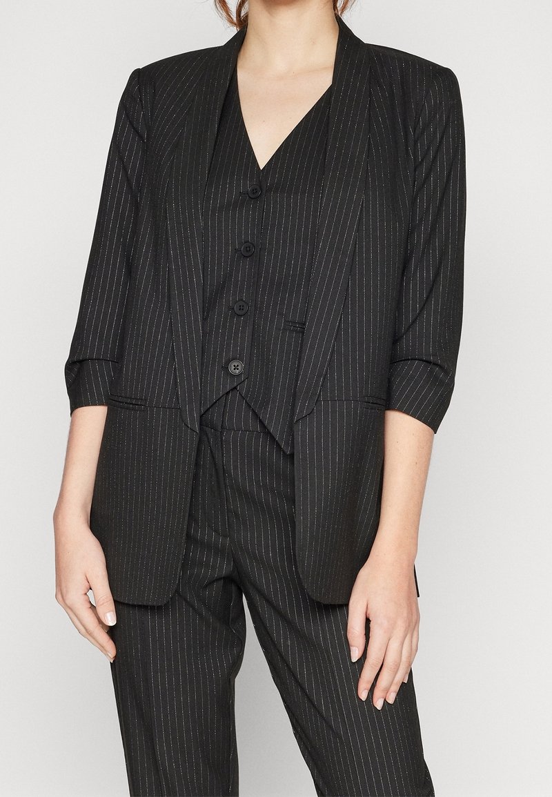 Femme portant un costume noir à fines rayures avec un gilet trois boutons, un blazer assorti à manches trois-quarts et un pantalon sur mesure.