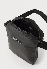 Černá nylonová crossbody taška obdélníkového tvaru, se zipovým uzávěrem a vnějším logem BOSS. Obsahuje nastavitelný popruh a měkkou podšívku.