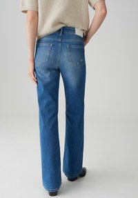 Jean en denim bleu bootcut avec un léger délavage, deux poches arrière et une taille de couleur claire. Porté avec un pull beige et duveteux.