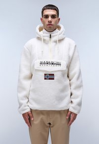 Pullover bianco in pelliccia con zip a metà, cappuccio con lacci, caratterizzato da una patch del logo frontale e un accento con la bandiera norvegese. Abbinato a pantaloni beige.
