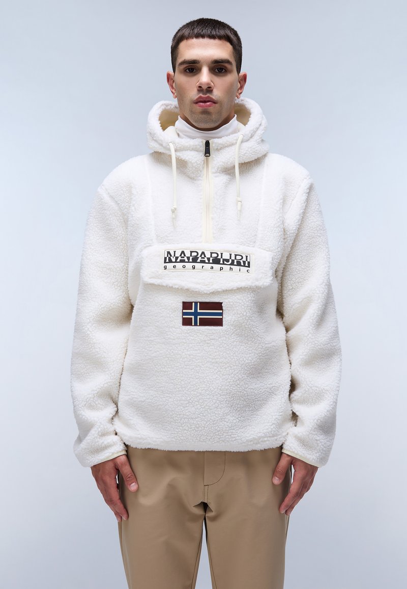 Pullover bianco in pelliccia con zip a metà, cappuccio con lacci, caratterizzato da una patch del logo frontale e un accento con la bandiera norvegese. Abbinato a pantaloni beige.