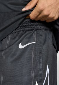 Čierne športové šortky z priedušného materiálu s bielym logom Nike, elastickým pásom a jemným vertikálnym prúžkovaním.