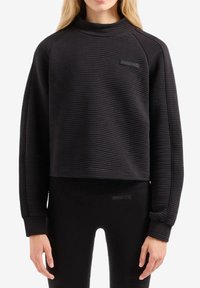Felpa cropped nera con texture a costine, colletto alto, maniche raglan e un piccolo logo sul petto. Abbinata a leggings neri.