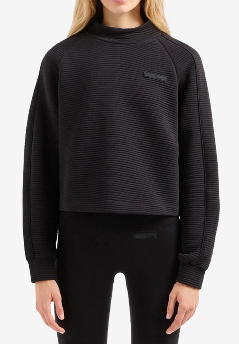 Felpa cropped nera con texture a costine, colletto alto, maniche raglan e un piccolo logo sul petto. Abbinata a leggings neri.