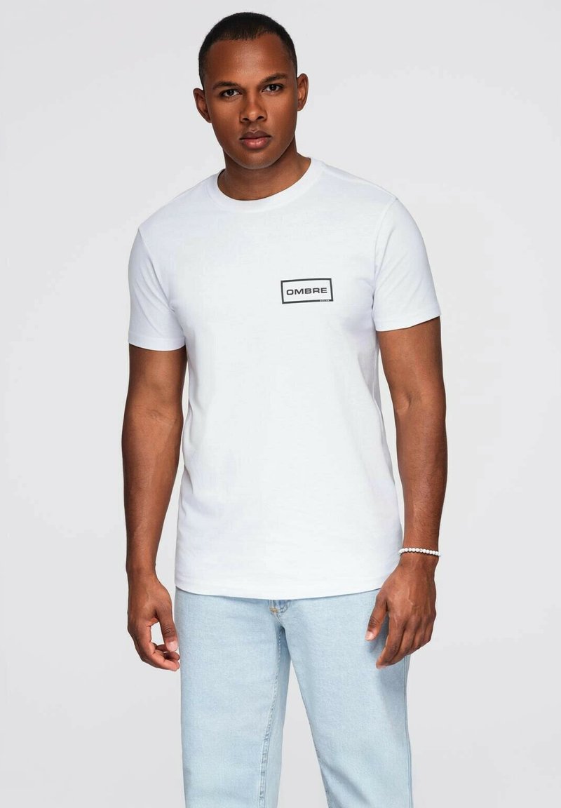 Mand iført en hvid kortærmet t-shirt med "OMBRE" logo på brystet og lyseblå jeans, som står mod en lys grå baggrund.