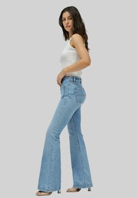 Jeans acampanados de color azul claro con una cintura alta, hechos de tela de mezclilla; cuentan con bolsillos traseros y un borde dobladillo. Combinados con una camiseta sin mangas blanca.