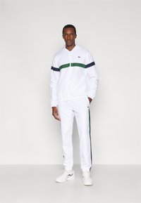 Fato de treino branco com riscas horizontais verdes e azul-marinho, composto por uma camisola com fecho e punhos elásticos. Acompanhado por sapatilhas desportivas brancas.