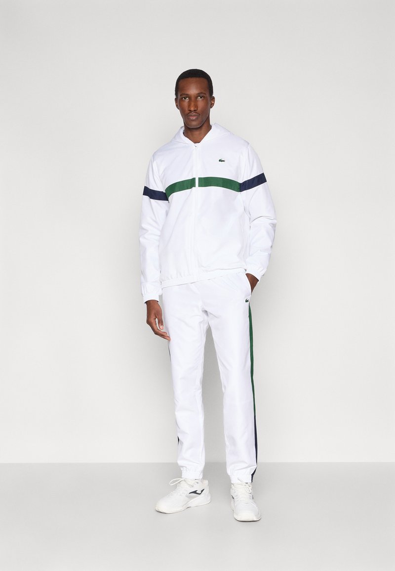 Fato de treino branco com riscas horizontais verdes e azul-marinho, composto por uma camisola com fecho e punhos elásticos. Acompanhado por sapatilhas desportivas brancas.