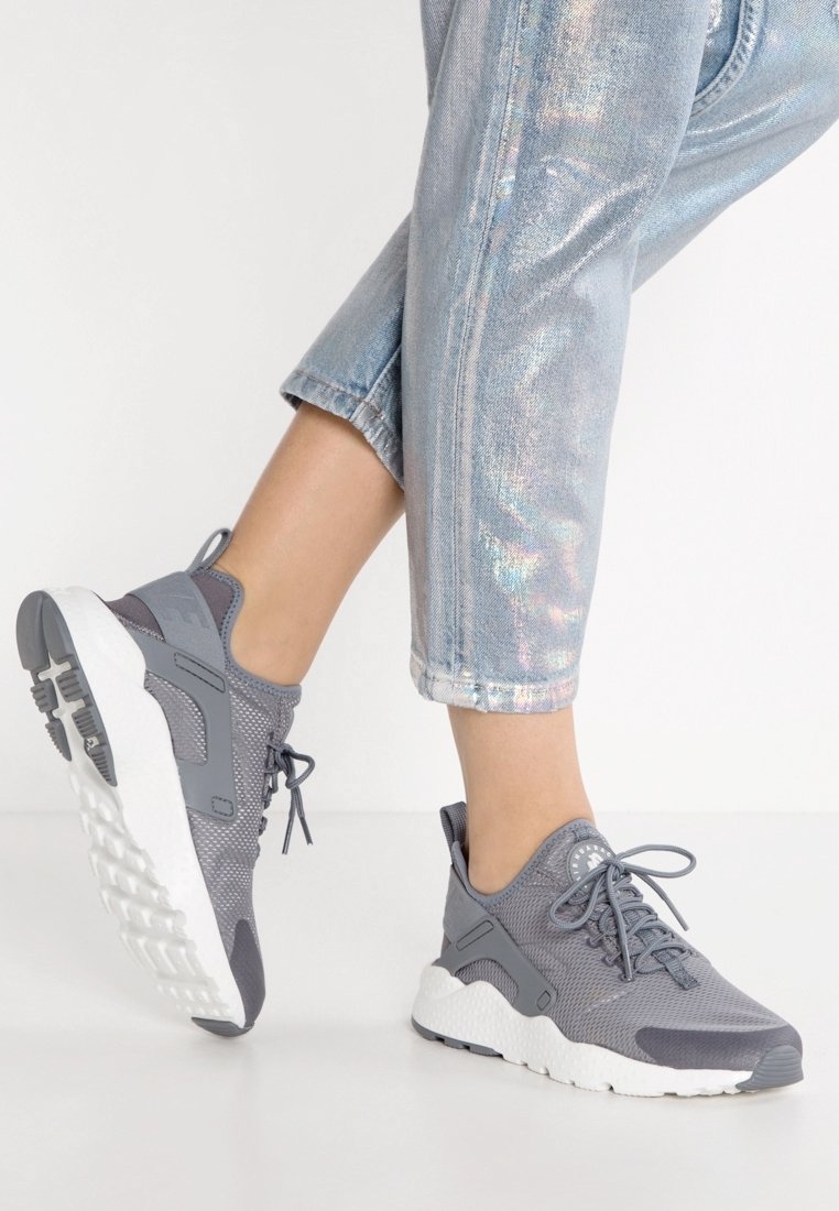 Personne portant un jean bleu clair brillant et des baskets de sport grises à semelles blanches, posant avec un pied levé.
