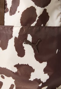 Tissu imprimé vache marron et blanc ; texture lisse ; logo Puma marron ; comprend des sangles marron ; détails de couture visibles.