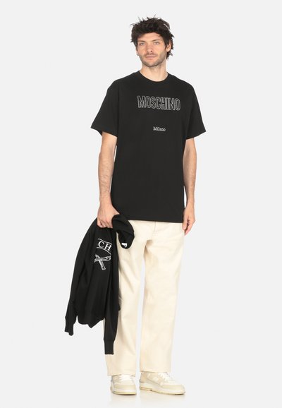 T-shirt in cotone nero con logo "MOSCHINO" in bianco. Vestibilità ampia, maniche corte, il modello tiene una felpa nera. Pantaloni chiari e sneakers.