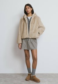 Chaqueta de borrego sintético beige con capucha, suéter gris debajo, minifalda a cuadros, calcetas grises y botines beige con costuras decorativas.