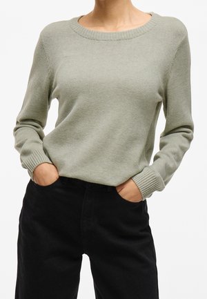 Femme portant un pull gris clair à col rond avec manches longues et pantalon noir, mains dans les poches sur fond blanc.