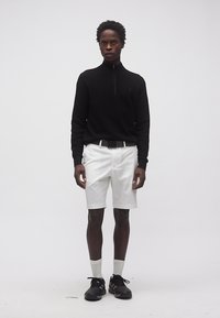 Άνδρας που στέκεται φορώντας μαύρο πουλόβερ με μακριά μανίκια, λευκή ζώνη και σορτς, λευκές κάλτσες και μαύρα αθλητικά παπούτσια σε απλό λευκό φόντο.