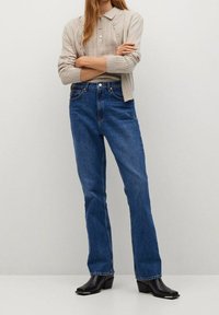 Siniset farkut suoralla lahkeella, joissa on klassinen viisipokkainen tyyli ja metallinen nappisuljenta, yhdistetty beigen väriseen neulepuseroon.