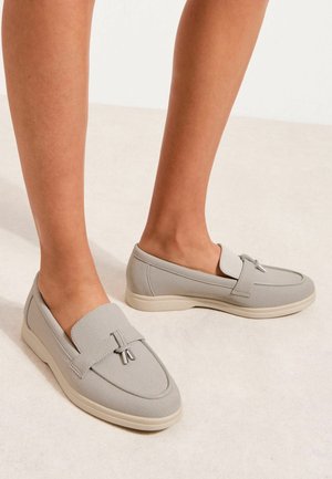 Fødder iført lysegrå slip-on loafers med runde tæer og dekorative kvaster på en lys neutral gulv.