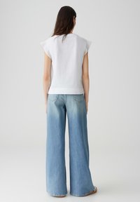 Witte mouwloze top met kantafwerking, gecombineerd met lichtblauwe wijdlopende jeans. Zacht materiaal, losse pasvorm en een subtiele vervaging in de jeans.