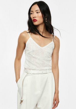 Donna con lunghi capelli neri, che indossa un top smanicato in pizzo bianco e pantaloni bianchi a vita alta con cintura abbinata, che guarda di lato.