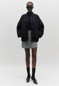 Zwarte oversized bomberjack, turtleneck, grijze minirok, zwarte kniehoge sokken, spitse hakken en donkere zonnebril. Strakke texturen.