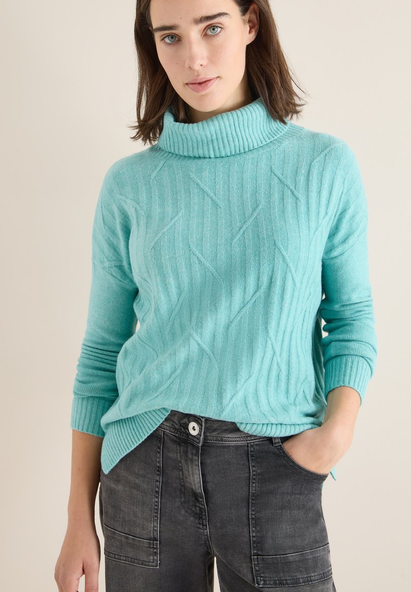 Pull en maille torsadée turquoise avec un col côtelé haut et des manches longues, associé à un pantalon cargo gris foncé avec des poches latérales.