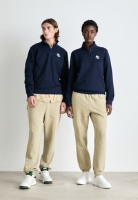 Jūras spēku zipsvārciņi ar Lacoste logo, beidge bikses un baltas kedas. Gluda auduma, brīva piegriezuma un apakšmalā ar elastīgu maliņu.