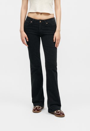 Γυναίκα με μαύρο τζιν bootcut, cropped λευκό αμάνικο τοπ και καφέ loafers με κρεμ αποχρώσεις, όρθια μπροστά σε λευκό φόντο.