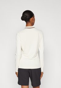 Žena s vlasy ve bunce, oblečená v bílé košili s dlouhými rukávy a pruhovaným límcem, a černých šortkách, obrácená zády k jednobarevnému pozadí.