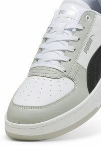 Puma-tennis kengät, joissa on valkoista nahkaa, harmaita yksityiskohtia ja musta logo. Teksturoidulla pinnalla on reikiä ja litteät nauhat.