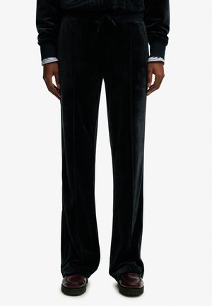 Zwarte fluwelen broek met wijde pijpen en zijzakken, gedragen met donkerrode loafers en een zwarte top met gestreepte mouwen zichtbaar bij de polsen.