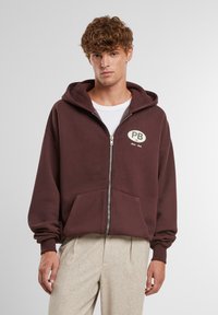 Bordová mikina na zips s prednou kapsou, voľný strih, kapucňa so šnúrkou a okrúhly vyšívaný logo "PB" na hrudi.
