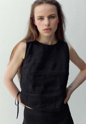Jonge vrouw met lang haar, draagend een mouwloos zwart topje, staand met handen in de zij tegen een effen lichte achtergrond.