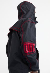 Chaqueta negra con detalles rojos, con capucha ajustable, gran solapa frontal y letras llamativas en la manga. Fabricada en un tejido ligero y texturizado.