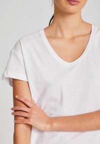 Tricou alb din bumbac, cu decolteu în V și mâneci scurte, răsucite, având o textura moale și o croială lejeră, vizibil parțial pe partea superioară a corpului unui model.