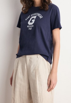 Nainen pukeutuneena laivansiniseen Gant Sportswear -t-paitaan ja beigenvärisiin laskostettuihin pöytiin, seisomassa vaalean yksivärisen seinän edessä.