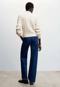 Kremowy sweter w warkoczowy splot z opadającymi ramionami, w połączeniu z ciemnoniebieskimi szerokimi spodniami i czarnymi butami na sznurowadła, widziany od tyłu.