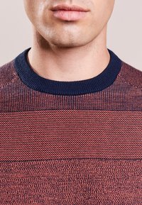 Rot- und marineblau gestreifter Strickpullover mit Rundhalsausschnitt und strukturiertem Stoff. Nahaufnahme der Nähte und Farbakzente.