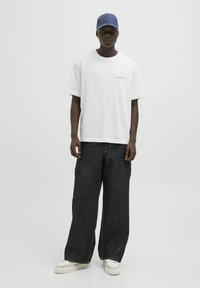 Camiseta de algodón blanca, de corte oversized, con un pequeño detalle en texto. Jeans de mezclilla oscuro de pierna ancha. Zapatillas blancas y una gorra azul completan el conjunto.