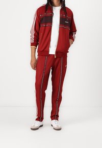 Röd träningsoverall med en zip-up jacka med en mörk horisontell rand och nitar, tillsammans med matchande byxor. Vit Adidas-logotyp och ränder.