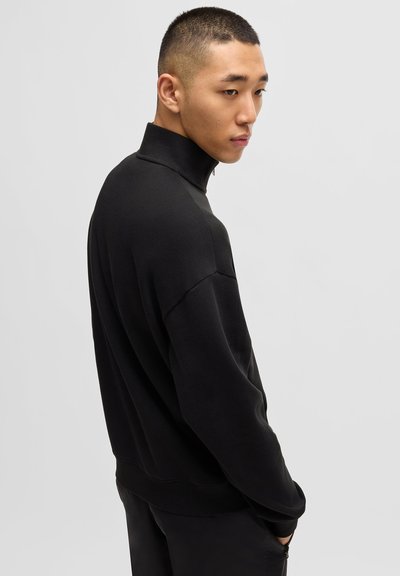 Sweatshirt noir à col montant, coupe ample, avec des poignets et un ourlet côtelés. Texture lisse et design minimaliste, mettant en avant une esthétique moderne et polyvalente.