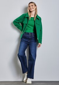 Grön texturerad jacka med två bröstfickor, bärs över en grön topp, i kombination med vida mörka jeans och vita sneakers.