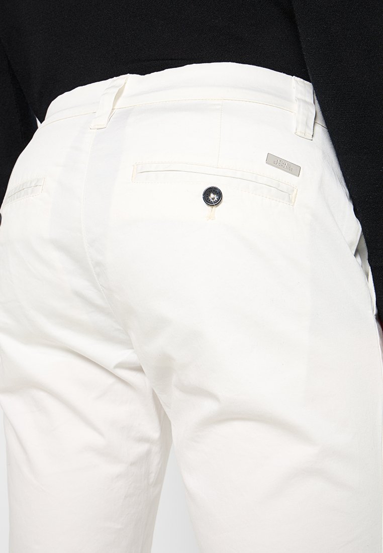 Pantaloni in cotone bianchi con una texture liscia, dotati di due tasche posteriori e chiusure con bottoni neri. Dettaglio di branding discreto vicino alla vita.