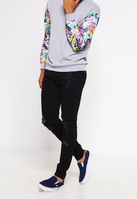 Sudadera gris con mangas de impresión gráfica colorida, combinada con jeans skinny negros desgastados y zapatos de lona azul marino.