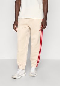 Beige Jogginghose mit zwei vertikalen roten Streifen entlang der Seiten. Mit gerippten Bündchen und einem roten Adidas-Logo auf der linken Seite.